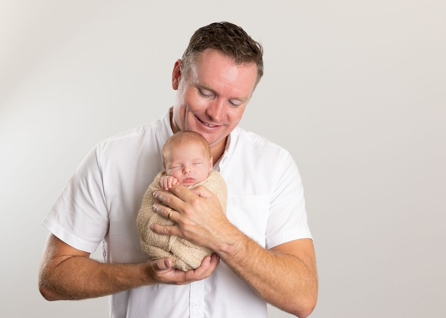 A smiling father in a white shirt tenderly holds his sleeping newborn baby wrapped in a beige swaddle.