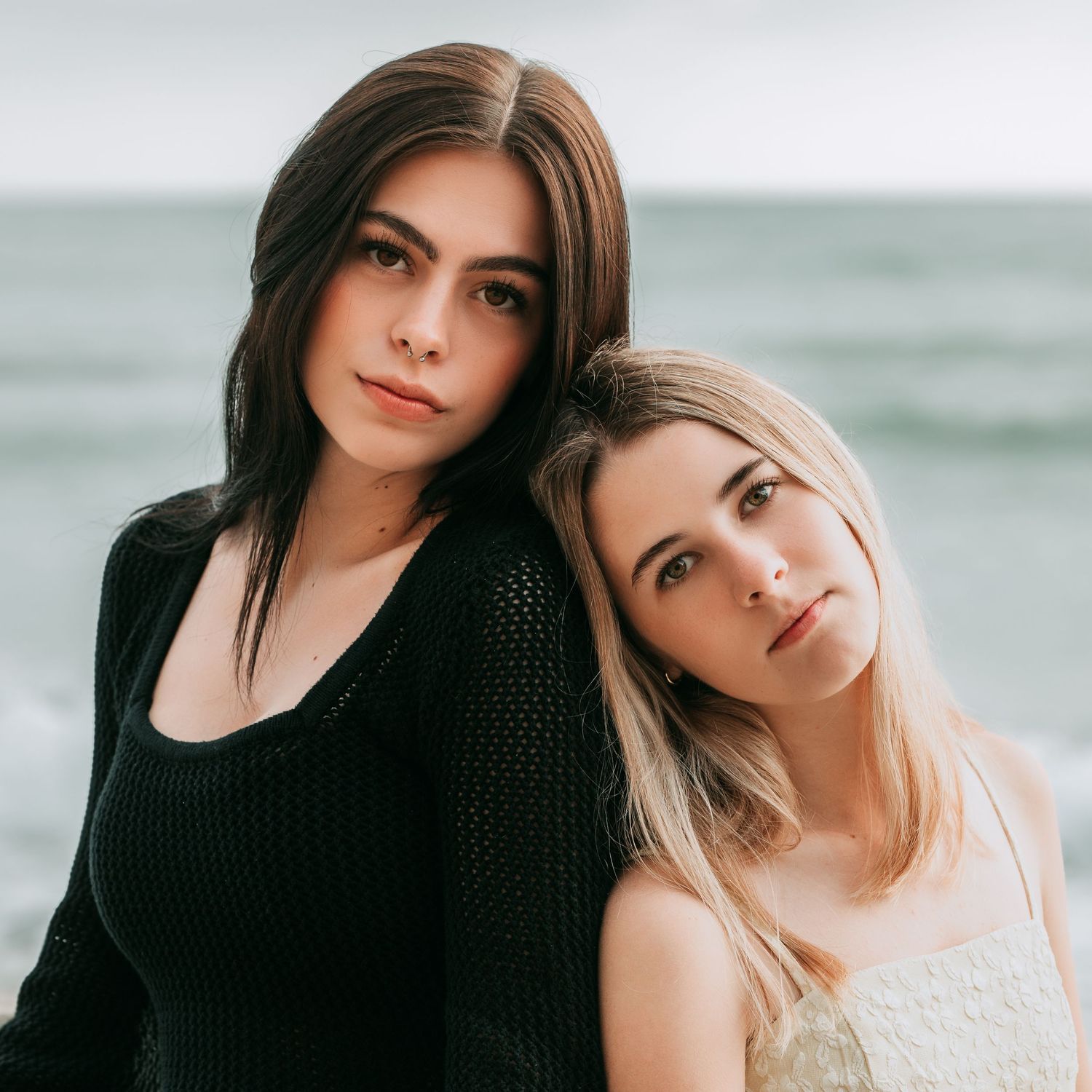 Two high school seniors in black and white outfits pose together at the beach with ocean waves in the background.