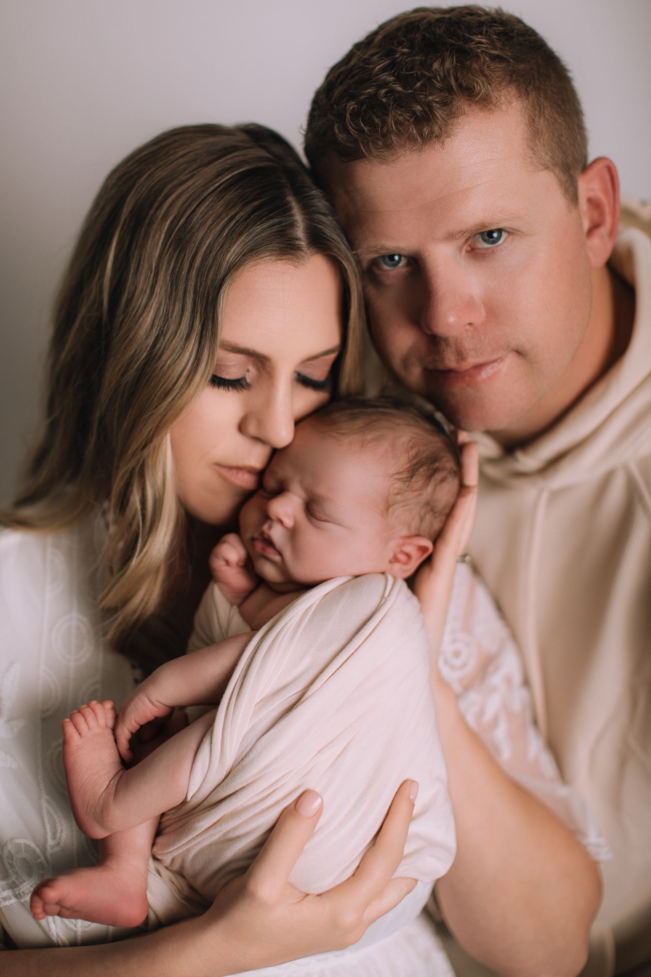 A couple tenderly holds their newborn baby, with the mother kissing the infant's head in a soft, warm portrait.