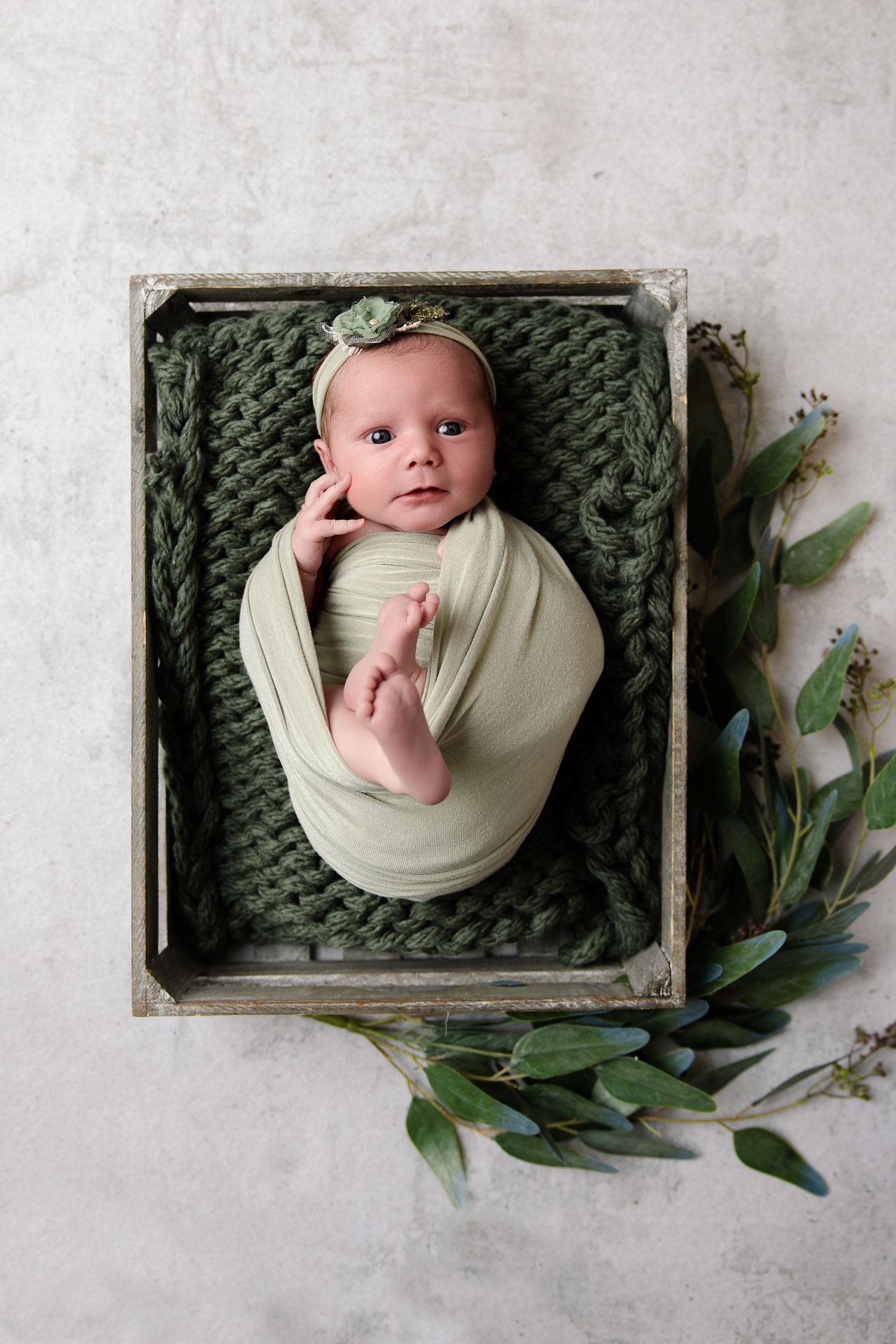 Newborn baby swaddled in green wrap lying in rustic wooden crate with knit blanket and floral headband.