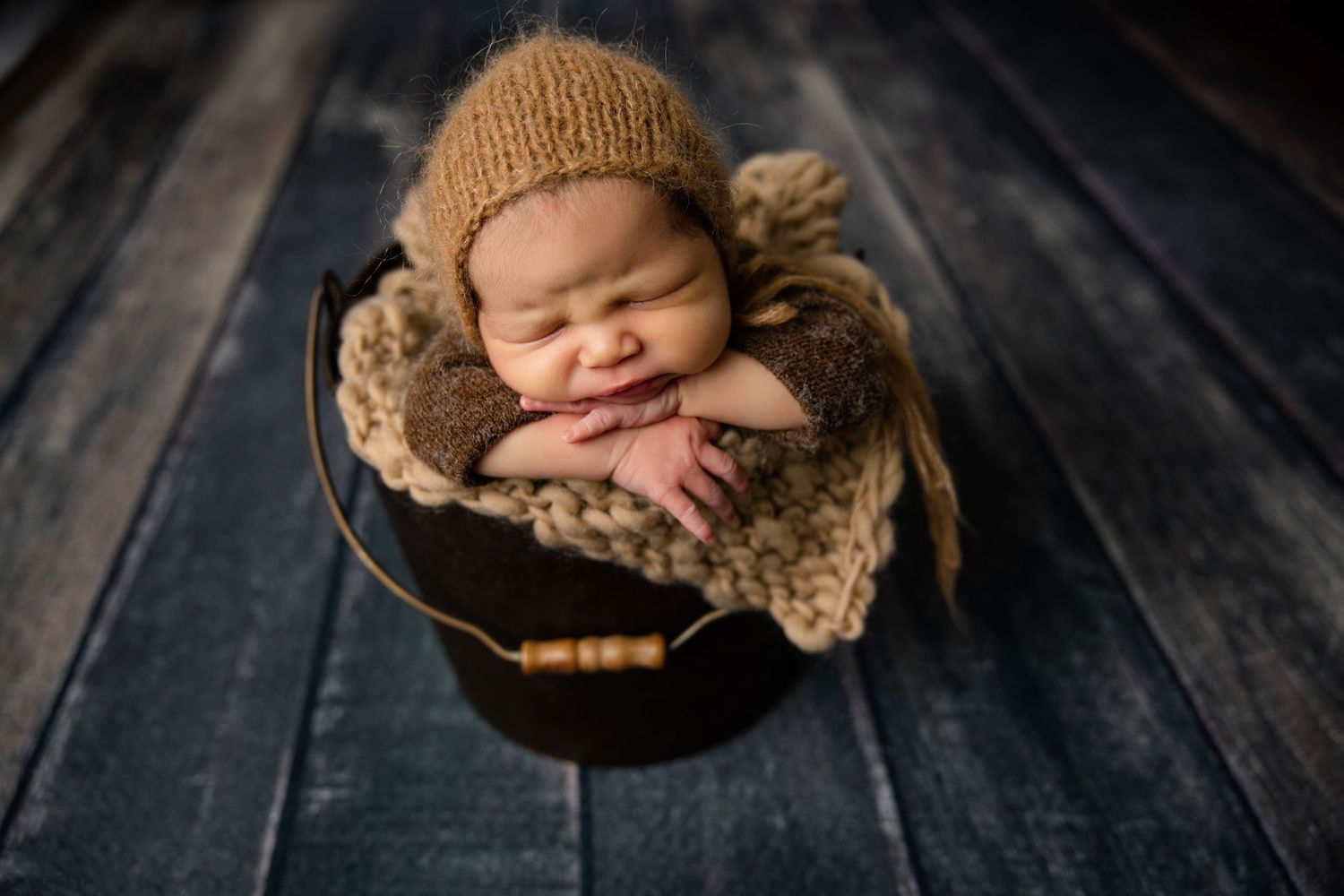 Smiling newborn baby in knit bonnet nestled in a dark bucket with cozy brown blankets on rustic wood floor.