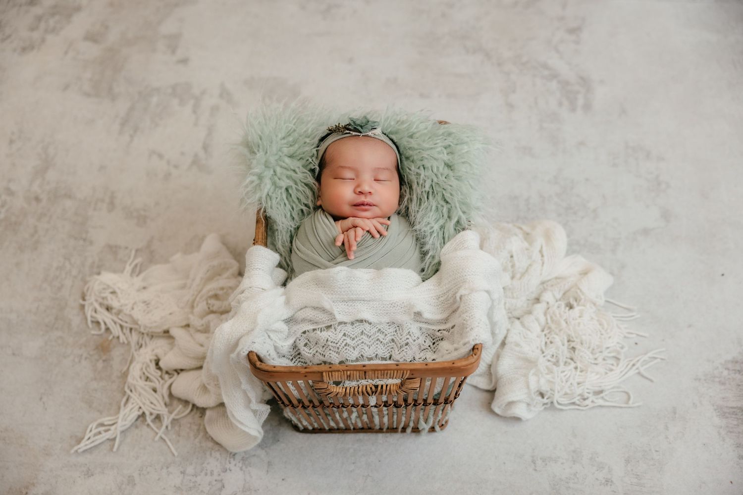 Newborn baby sleeping peacefully in a wicker basket with soft green fur and white wraps in a professional photo.