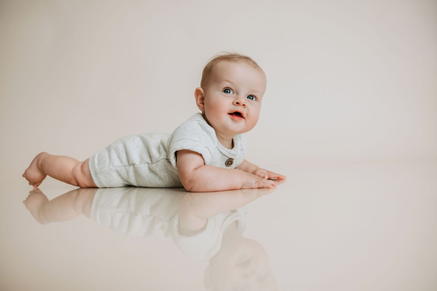 Adorable baby in a white onesie lying on a shiny floor, looking up curiously in a bright studio setting.