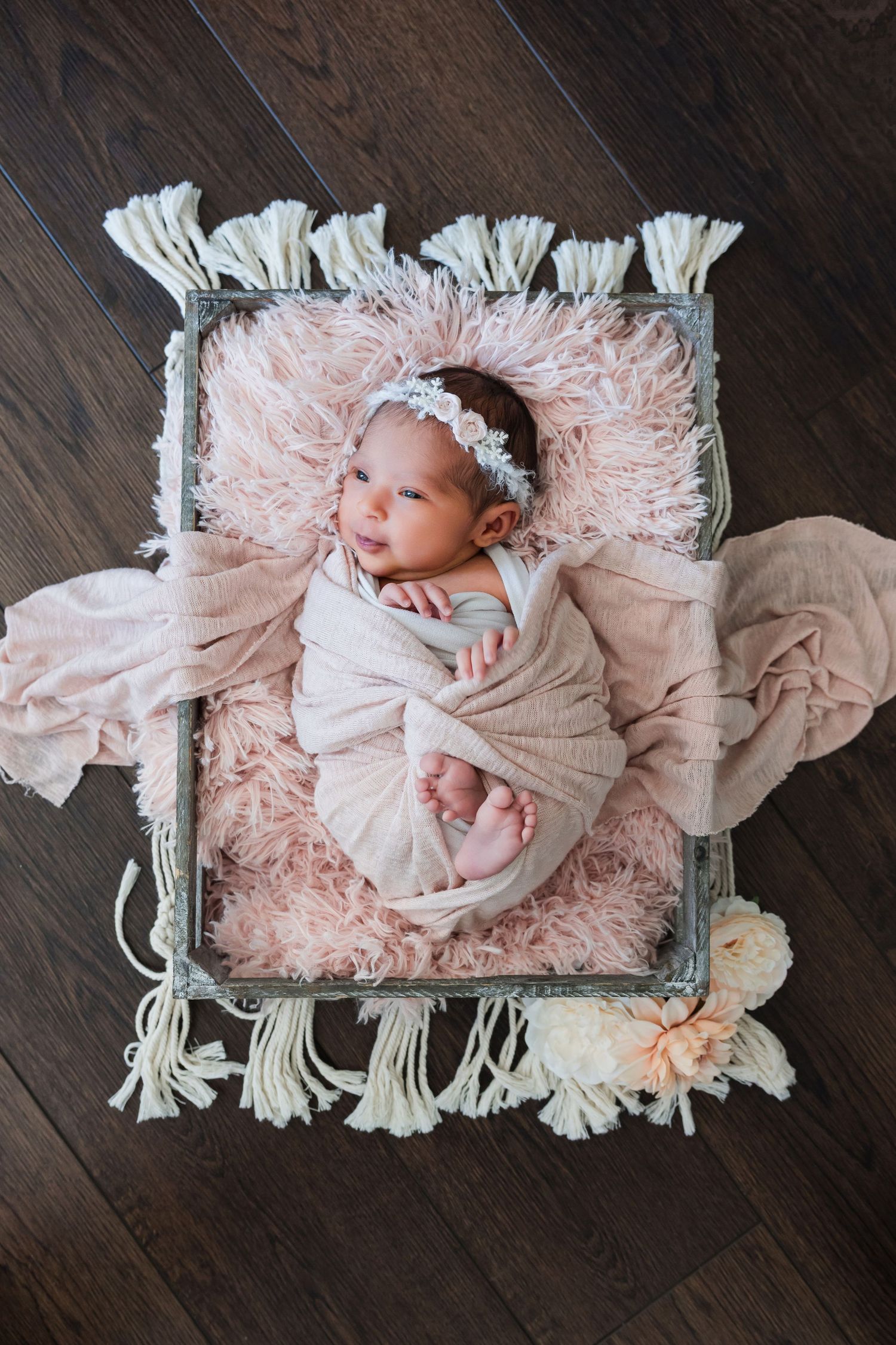 Newborn baby girl swaddled in pink wrap with floral headband, resting in a rustic crate with pink fur and tassels.