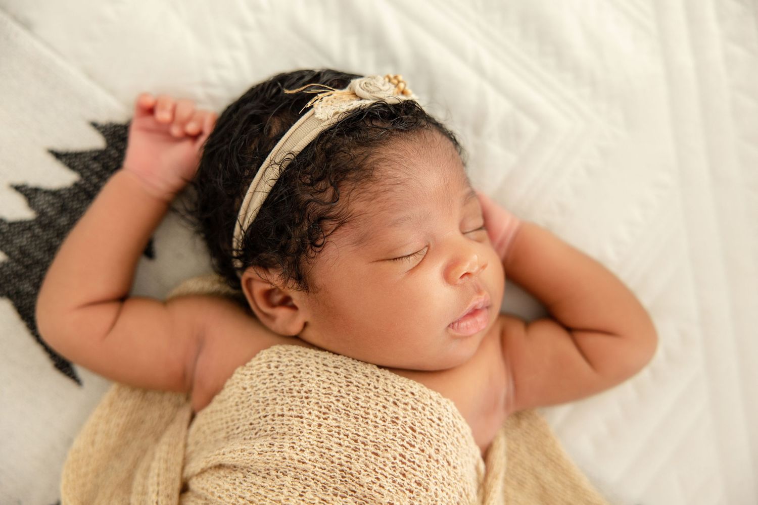 Peaceful newborn baby girl sleeping wrapped in a beige knit blanket wearing a delicate gold headband.