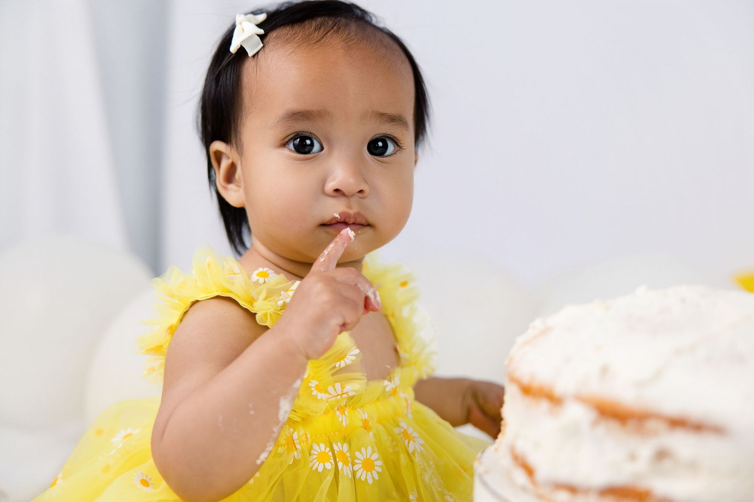Cute toddler girl in yellow dress tasting cake frosting with finger at birthday celebration.
