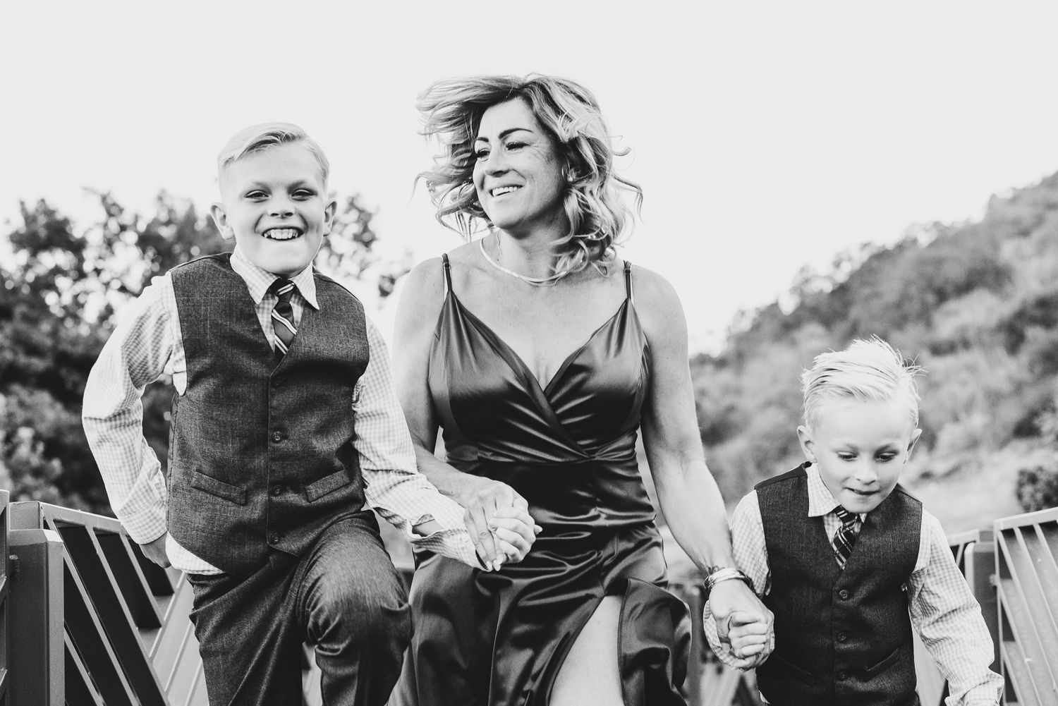 Black and white photo of smiling woman holding hands with two boys in dress vests outdoors.