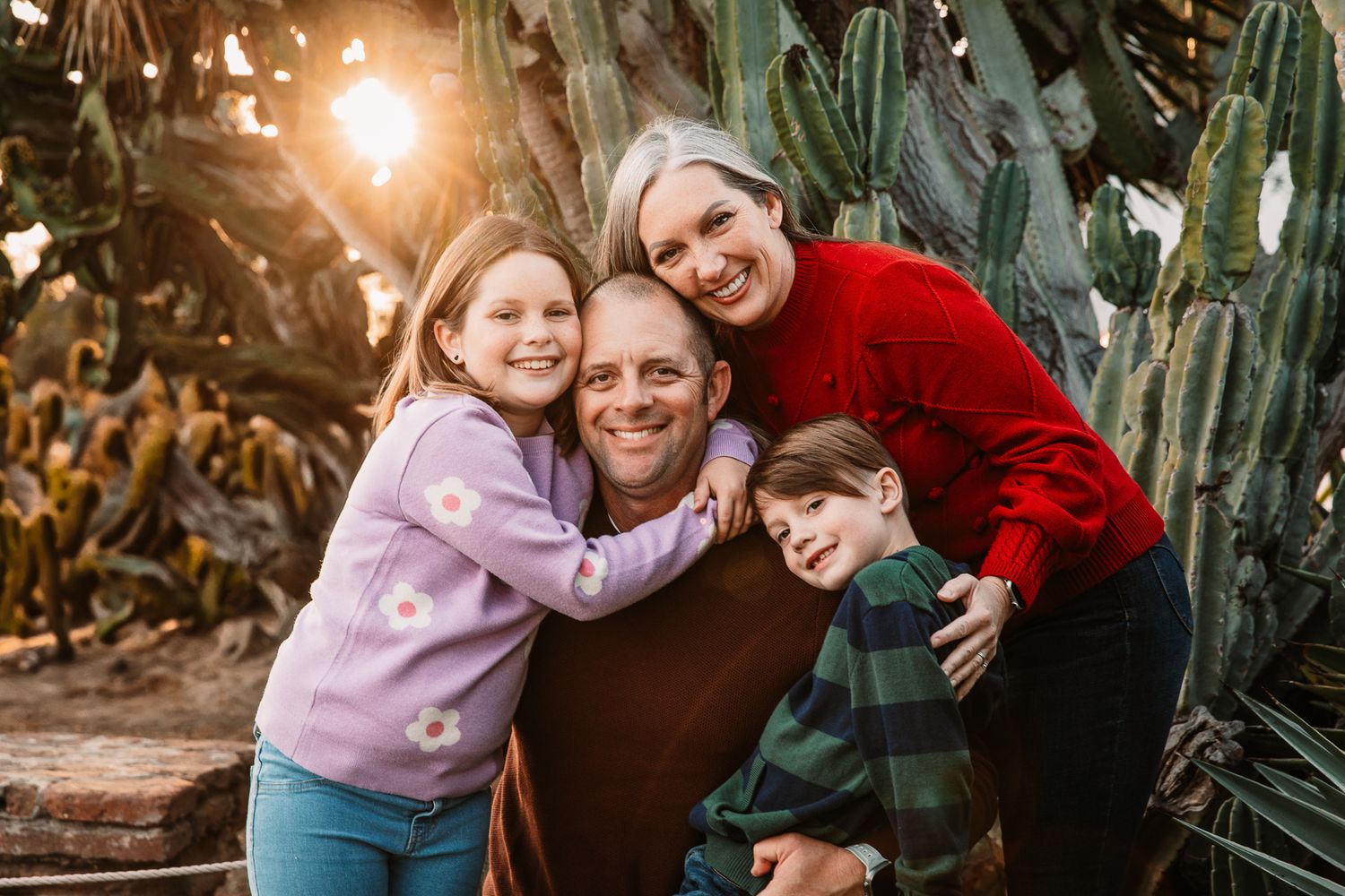 Happy family of four hugging and smiling together in front of tall cactus plants in warm sunlight in Oceanside, CA