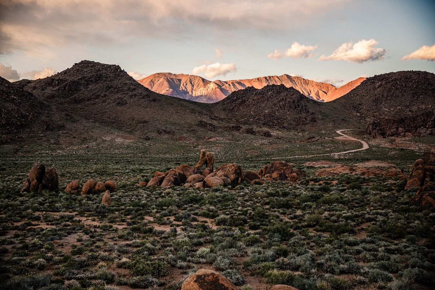 Desert landscape with rocky terrain, winding road, and snow-capped mountains at sunset in a vast wilderness.
