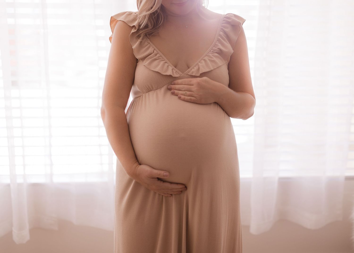 Pregnant woman in a blush ruffled dress gently cradling her baby bump near a softly lit window in Bonsall, near Fallbrook, & Temecula