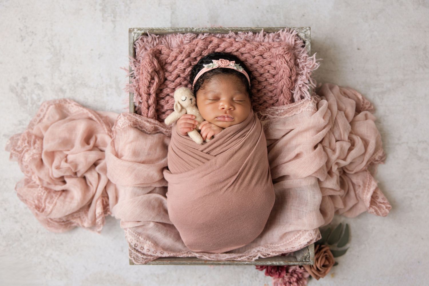 Sleeping newborn baby girl wrapped in pink swaddle with floral headband lying in wooden box for newborn photos.