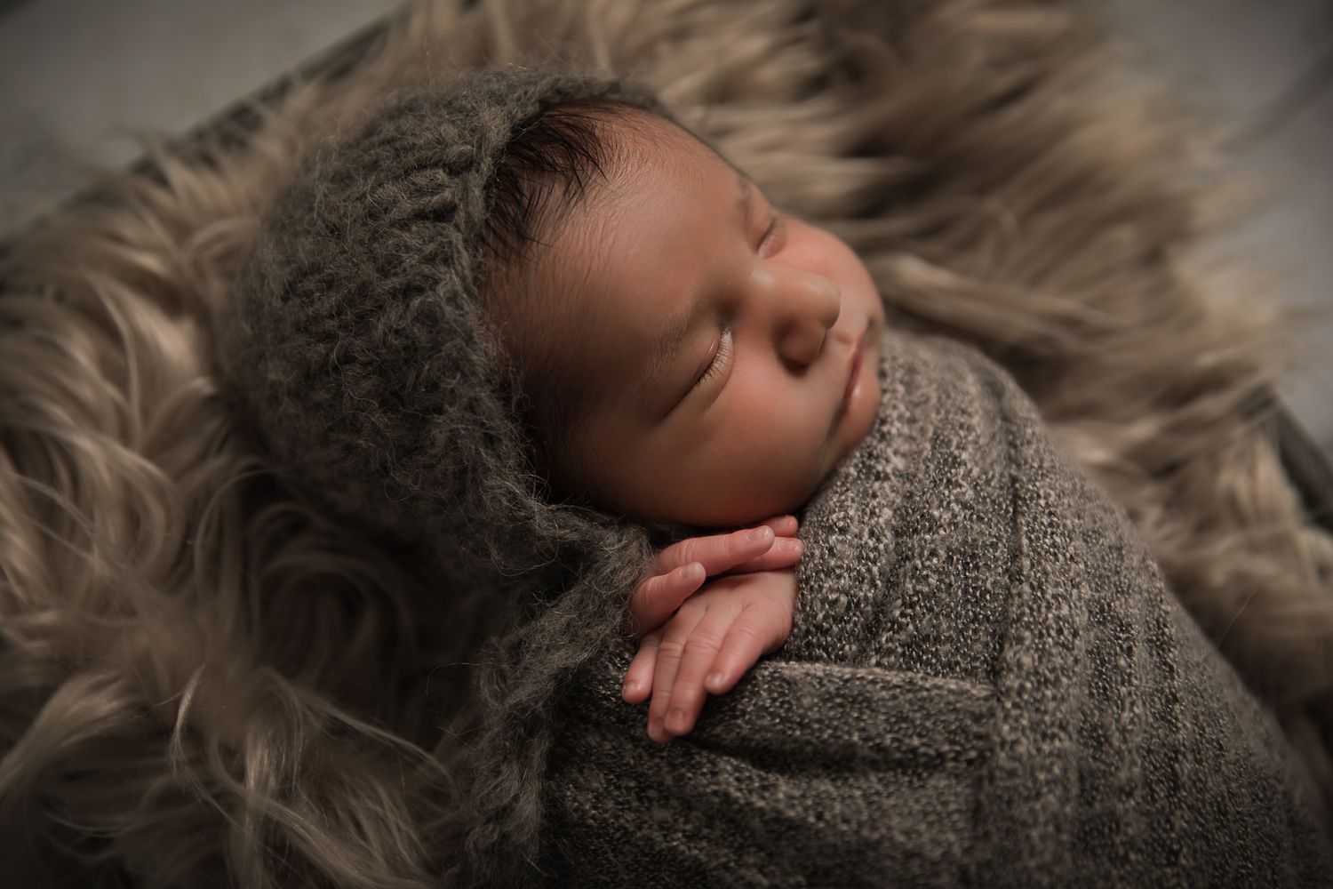 Sleeping newborn baby swaddled in gray knit wrap with matching bonnet on fur blanket.
