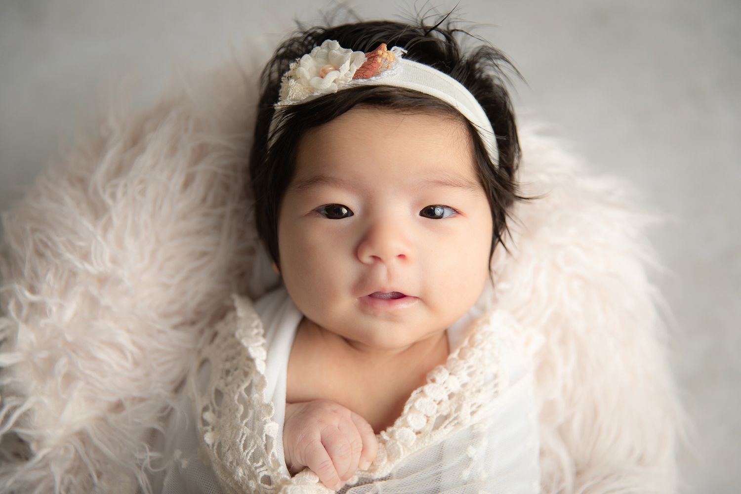 Alert newborn baby girl with dark hair wearing a floral headband looks up from a white fur wrap in Carlsbad, near Bonsall
