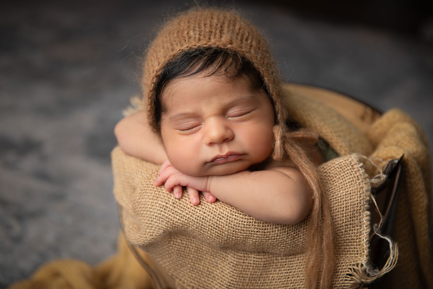 Sleeping newborn baby in burlap wrap wearing knit bonnet in rustic bowl. Fallbrook Newborn Photographer