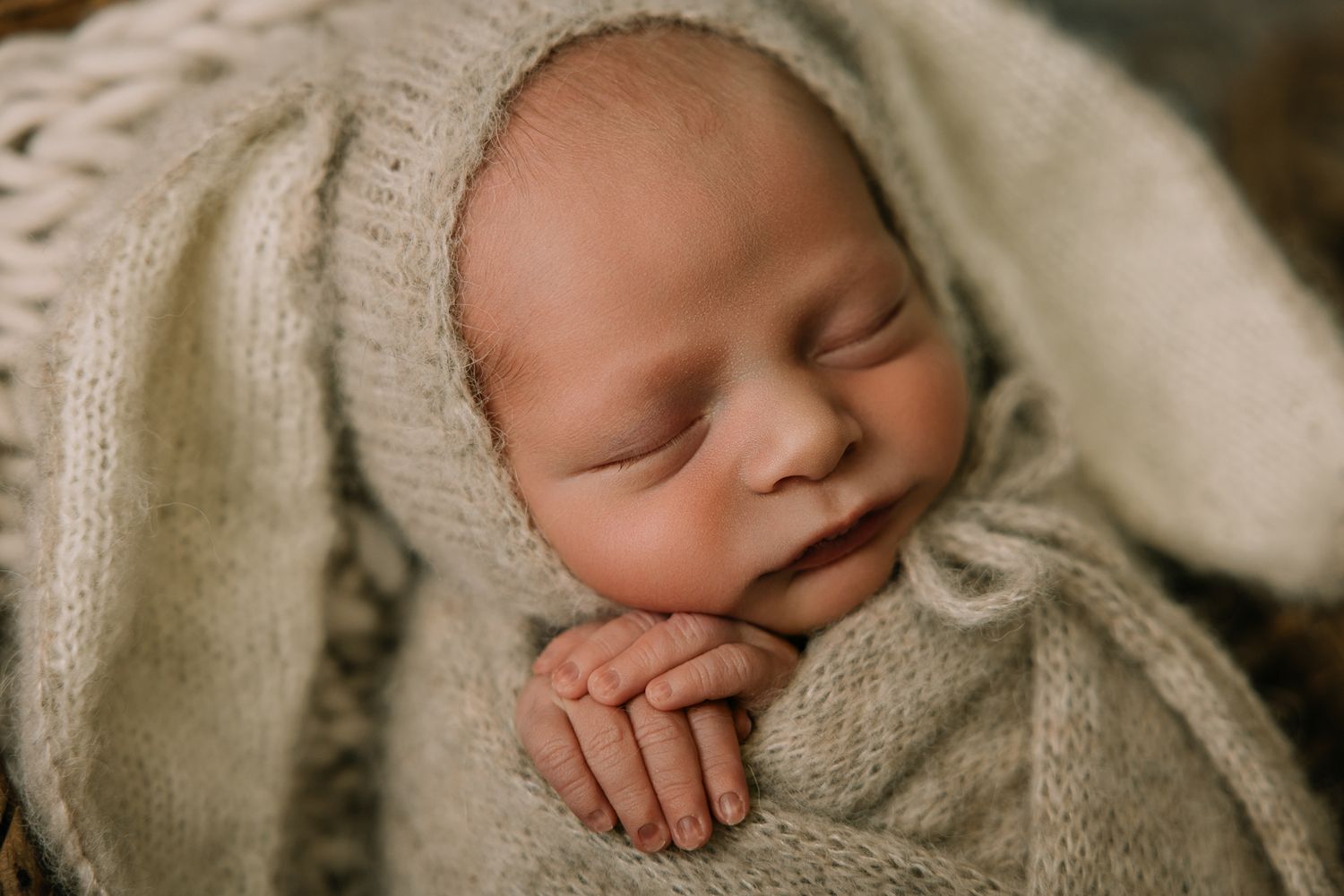Sleeping newborn baby in knit bonnet and wrap resting hands together in cozy basket.