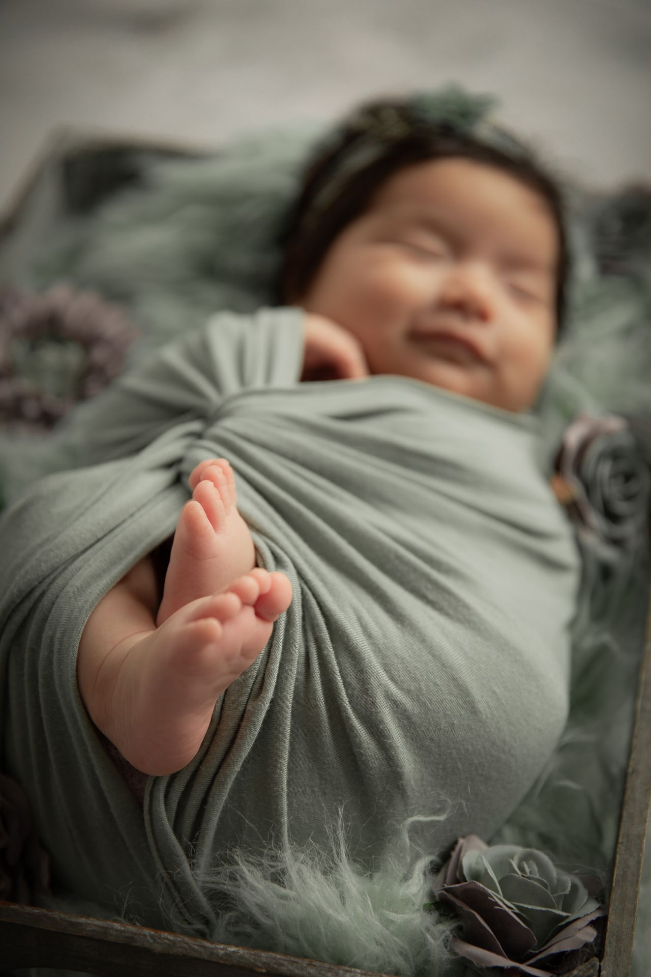 Smiling newborn baby wrapped in sage green swaddle with floral headband, tiny foot peeking out at The Studio Bonsall, near Fallbrook/Vista