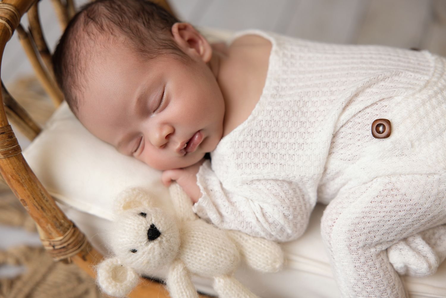 Peaceful newborn baby sleeping in a wicker bassinet wearing a white knit outfit, cuddling a small teddy bear.