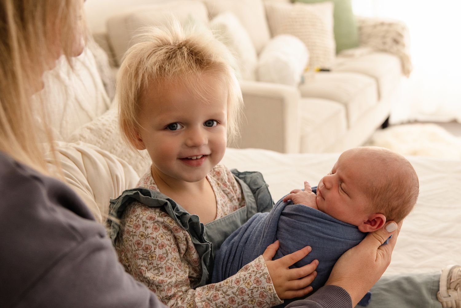 Toddler girl smiling while holding a swaddled newborn baby in a bright, cozy living room setting.