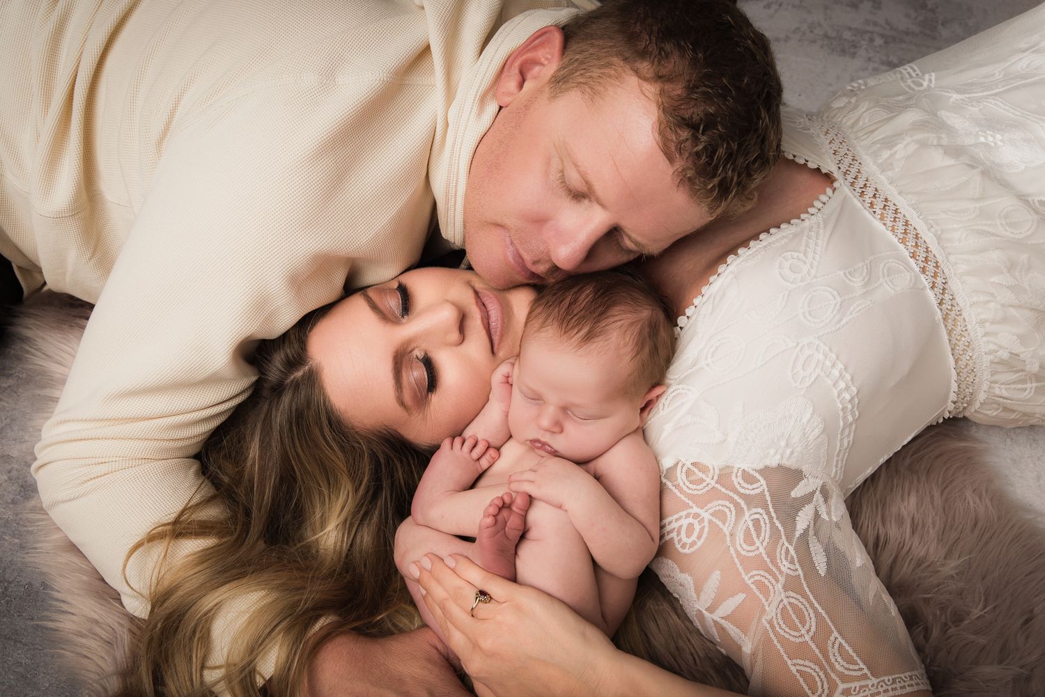 New parents lying down with their sleeping newborn baby between them in a tender family portrait.
