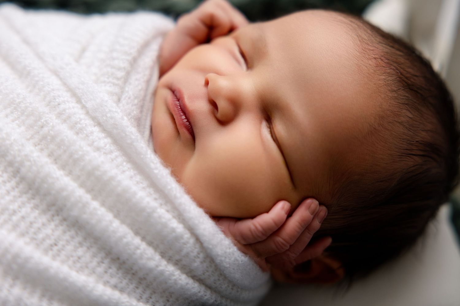 A peaceful newborn baby swaddled in a soft white blanket, sleeping with tiny hand tucked under chin.