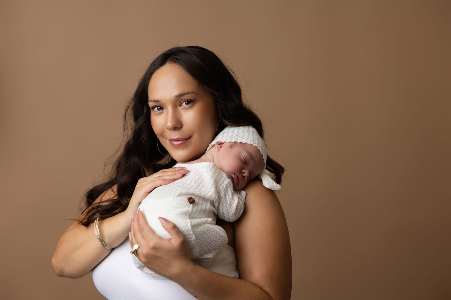 A mother in a white dress tenderly holds her sleeping newborn baby against a warm beige studio background.