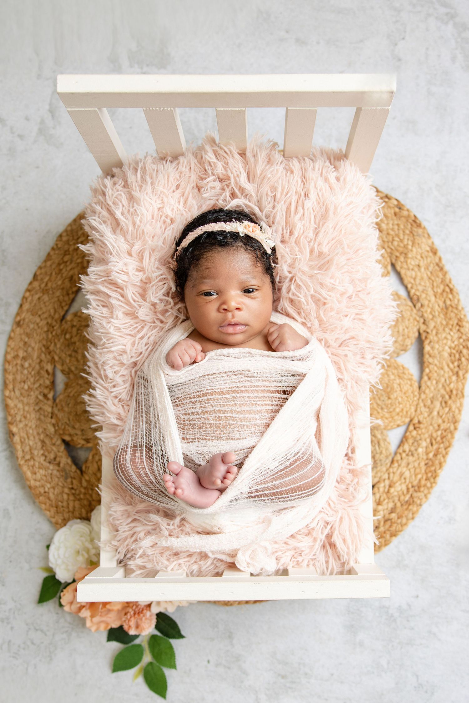 Newborn baby girl swaddled in pink wrap lying on fluffy pink fur in a tiny white bed with floral accents.