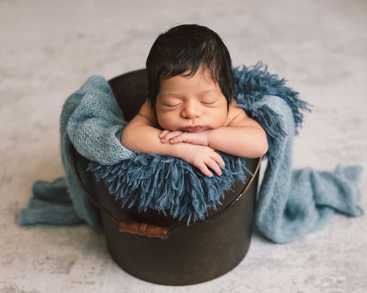 Sleeping newborn baby posed in rustic wooden bucket with teal blue blanket for professional photo.