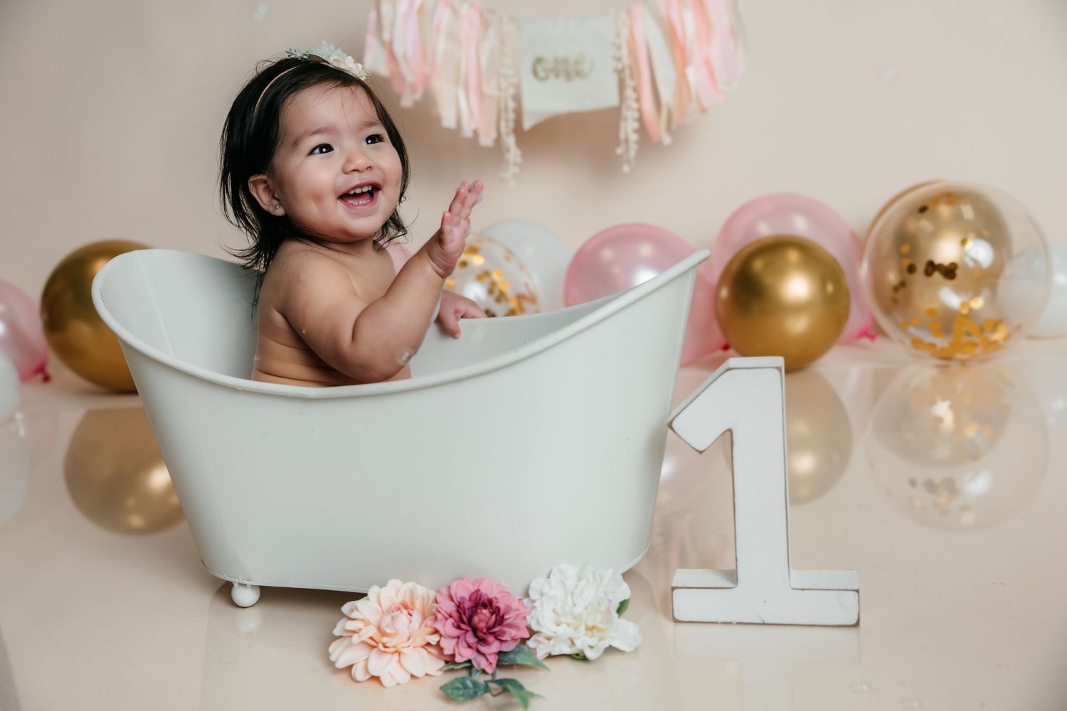 Smiling baby girl clapping hands in white mini bathtub with pink and gold balloons and number one sign.