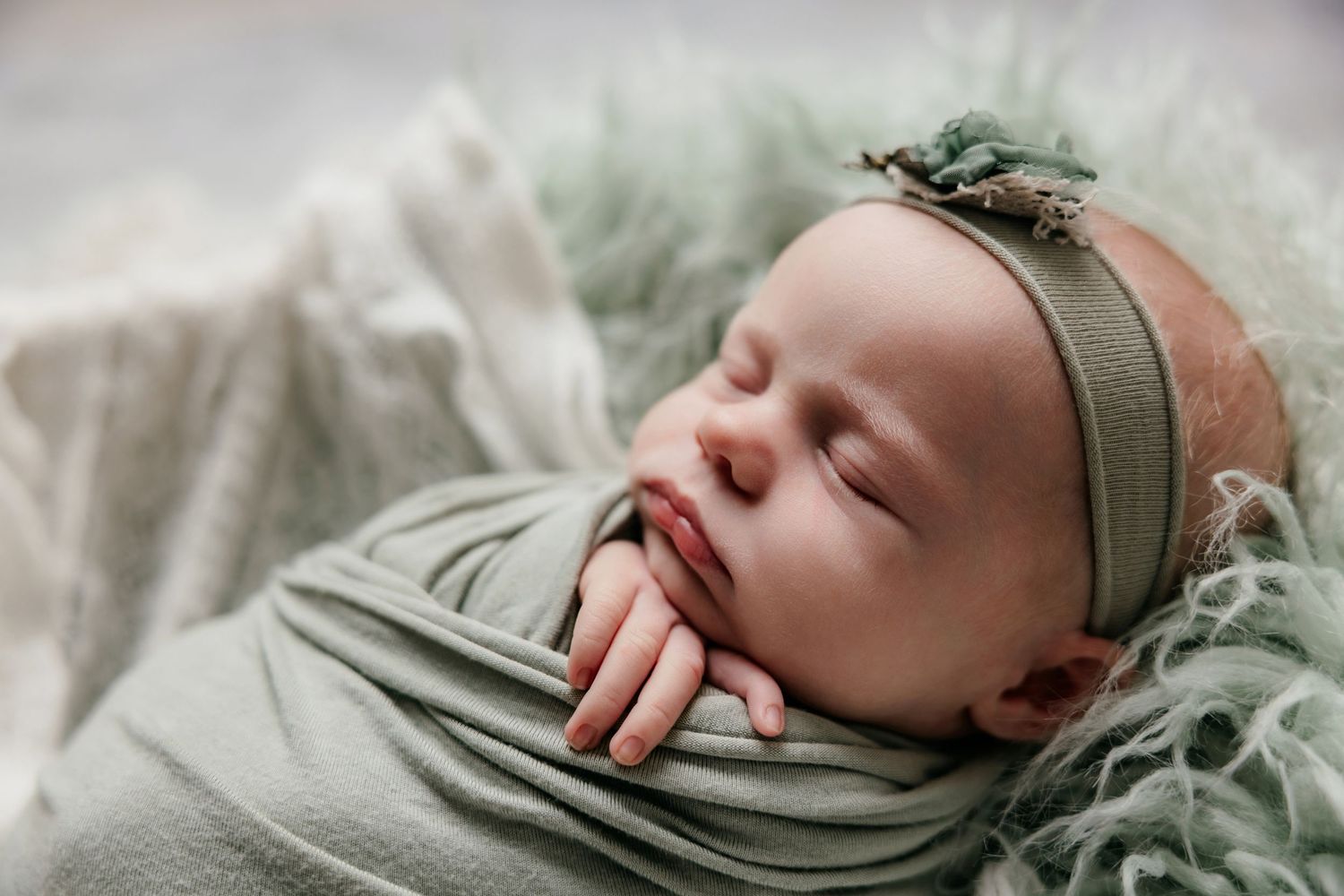 Peaceful newborn baby wrapped in sage green swaddle with floral headband, sleeping on soft green textured blanket.