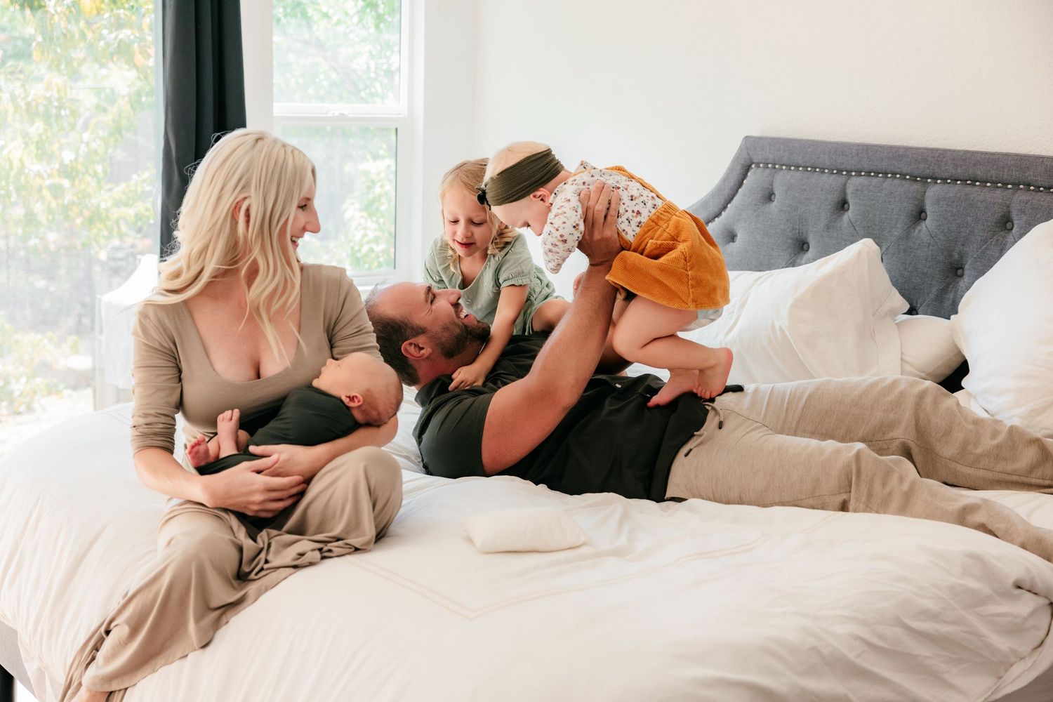 Happy family with two toddlers and newborn baby playing together on white bed at home.