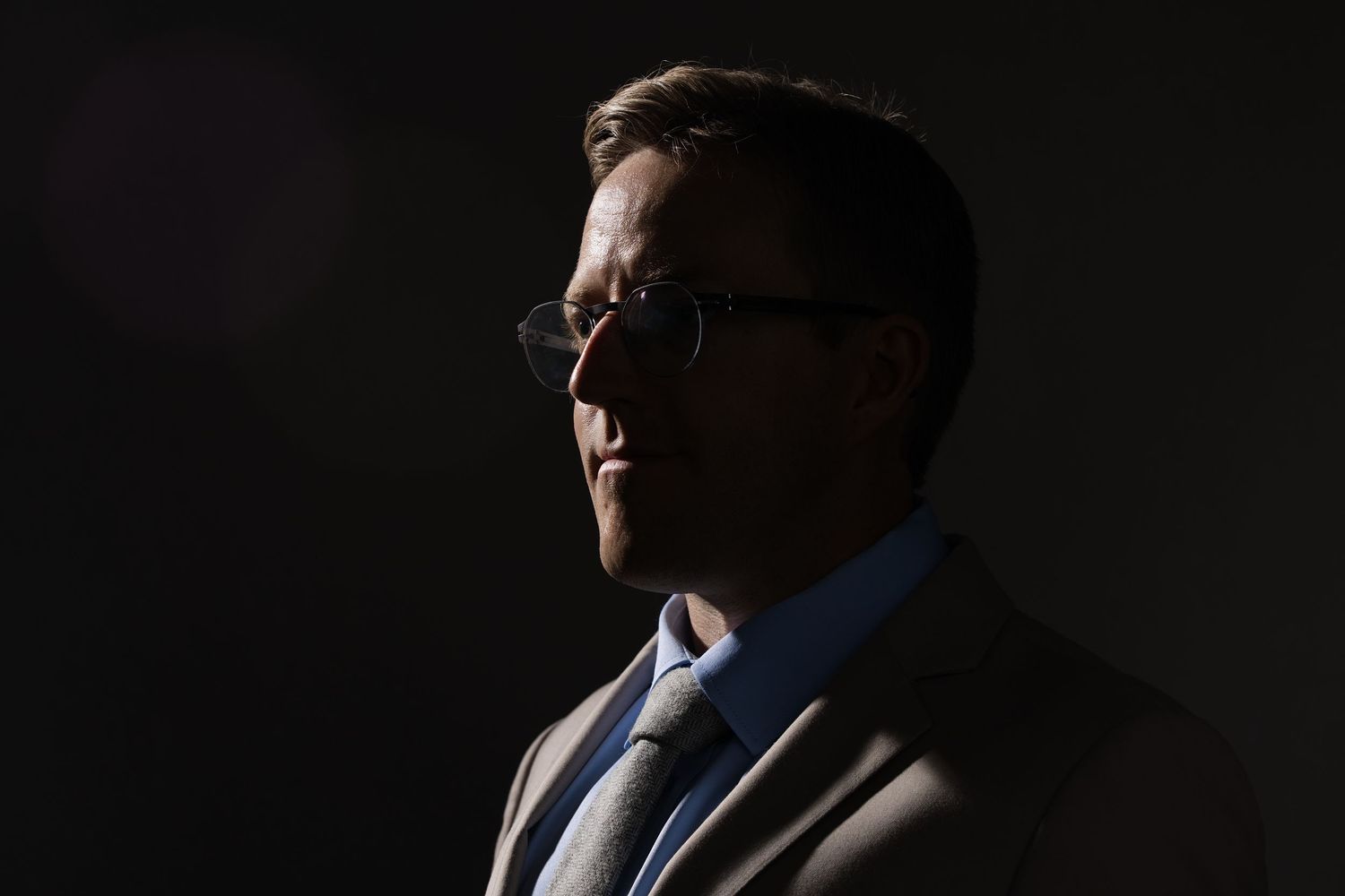 A man in a suit and glasses is dramatically lit against a dark background in a moody portrait photo.
