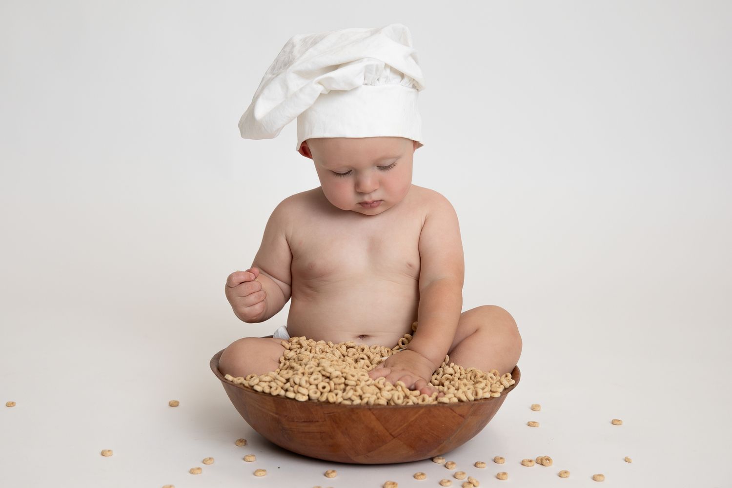 Baby wearing a white chef hat sitting in a wooden bowl filled with puffed grain cereal.