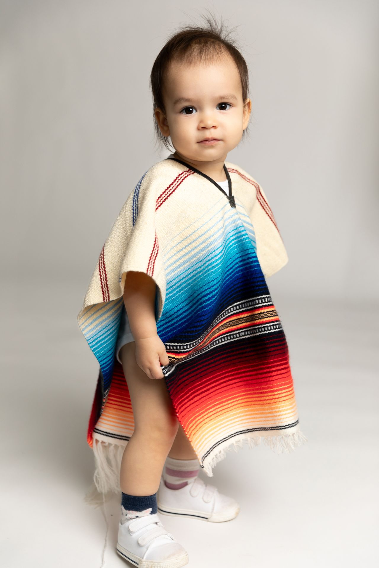 Adorable baby wearing a colorful serape poncho standing on a light gray background.