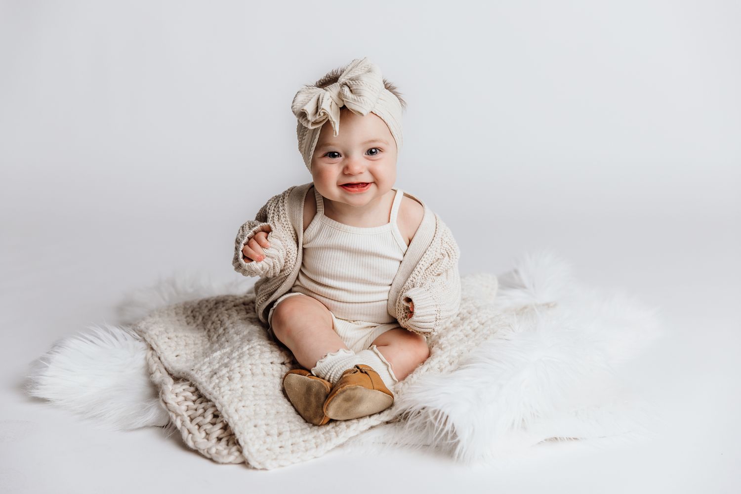 Adorable smiling baby in neutral outfit with bow headband sitting on knit blanket.