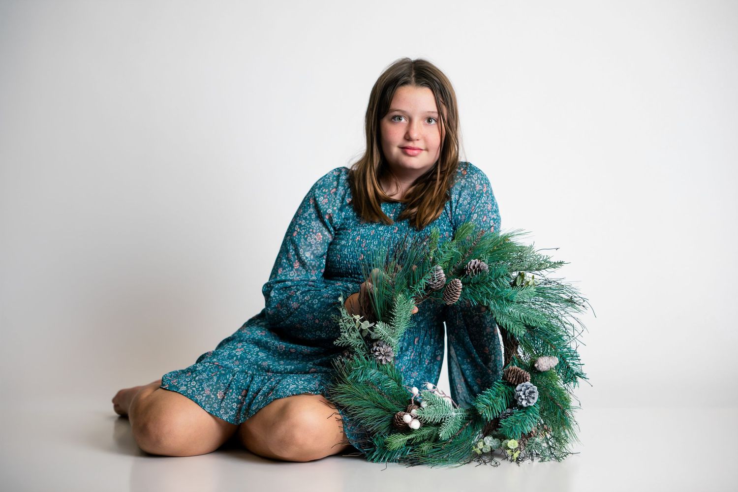 Young girl in teal floral dress sitting on white floor holding a festive Christmas wreath with pine cones.
