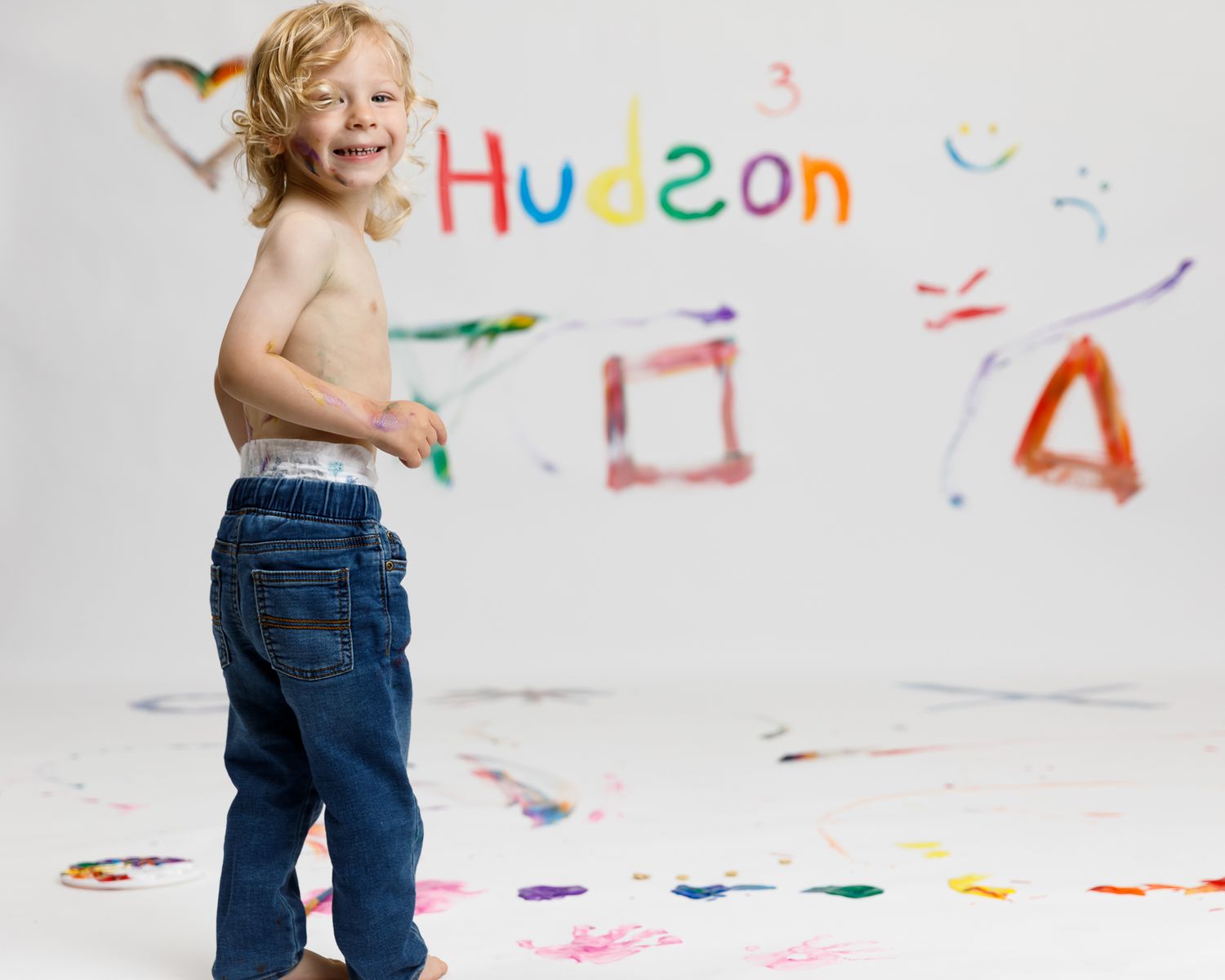 Smiling toddler boy named Hudson standing shirtless in jeans in front of colorful painted wall art.