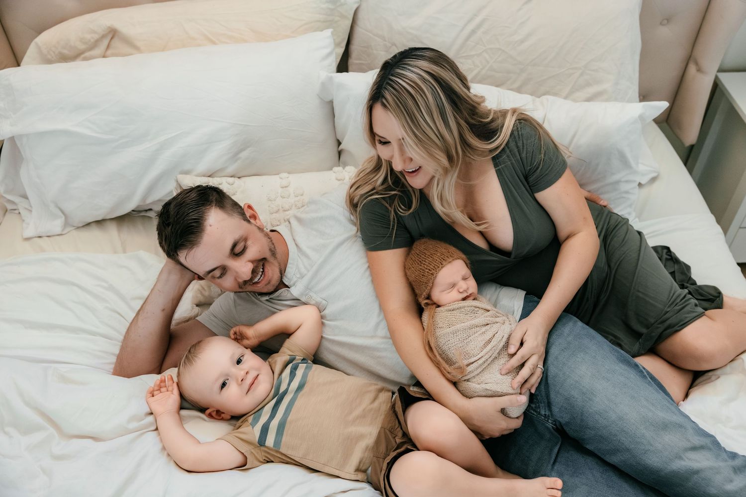 Happy family of four relaxing on a white bed, parents smiling at newborn baby while toddler plays nearby.