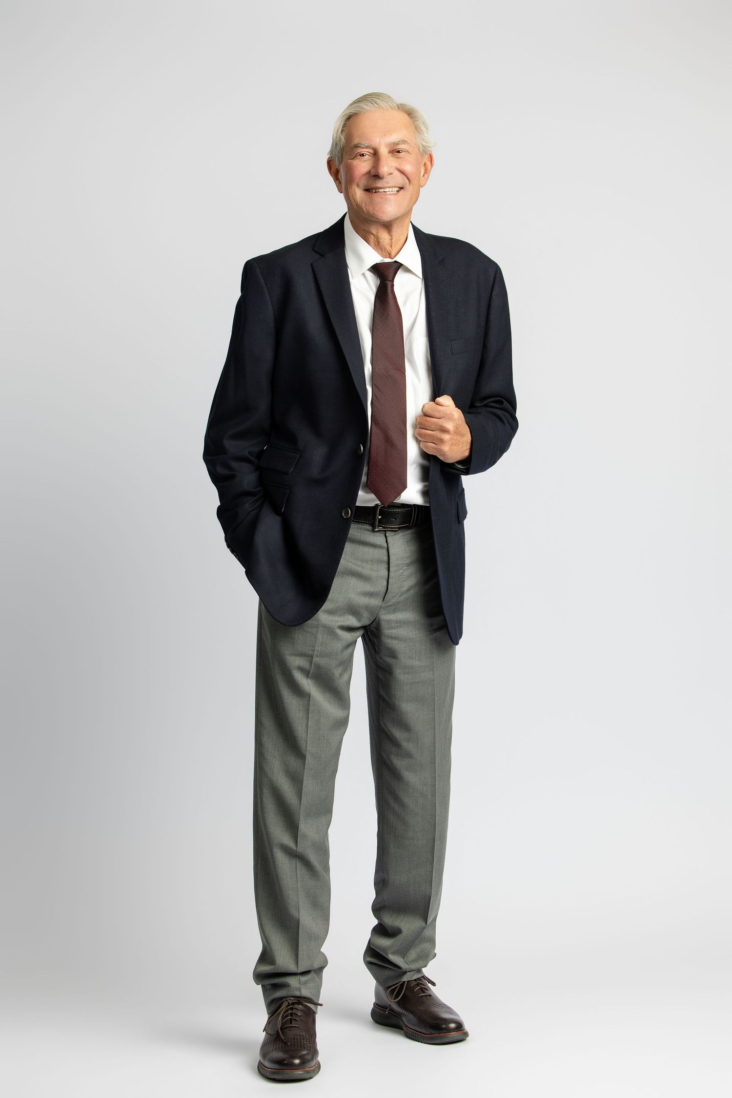 Smiling senior businessman in navy blazer, tie, and gray trousers standing confidently against a light background.