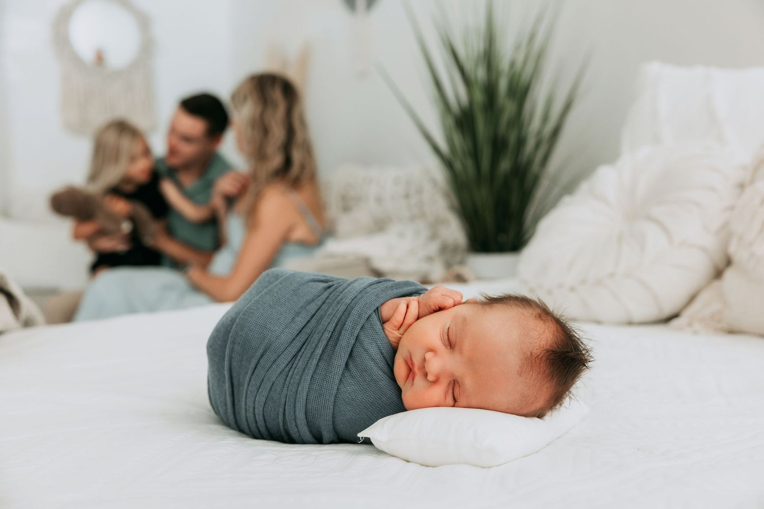 Newborn baby swaddled in gray wrap sleeping on white bed with family in soft-focus background.