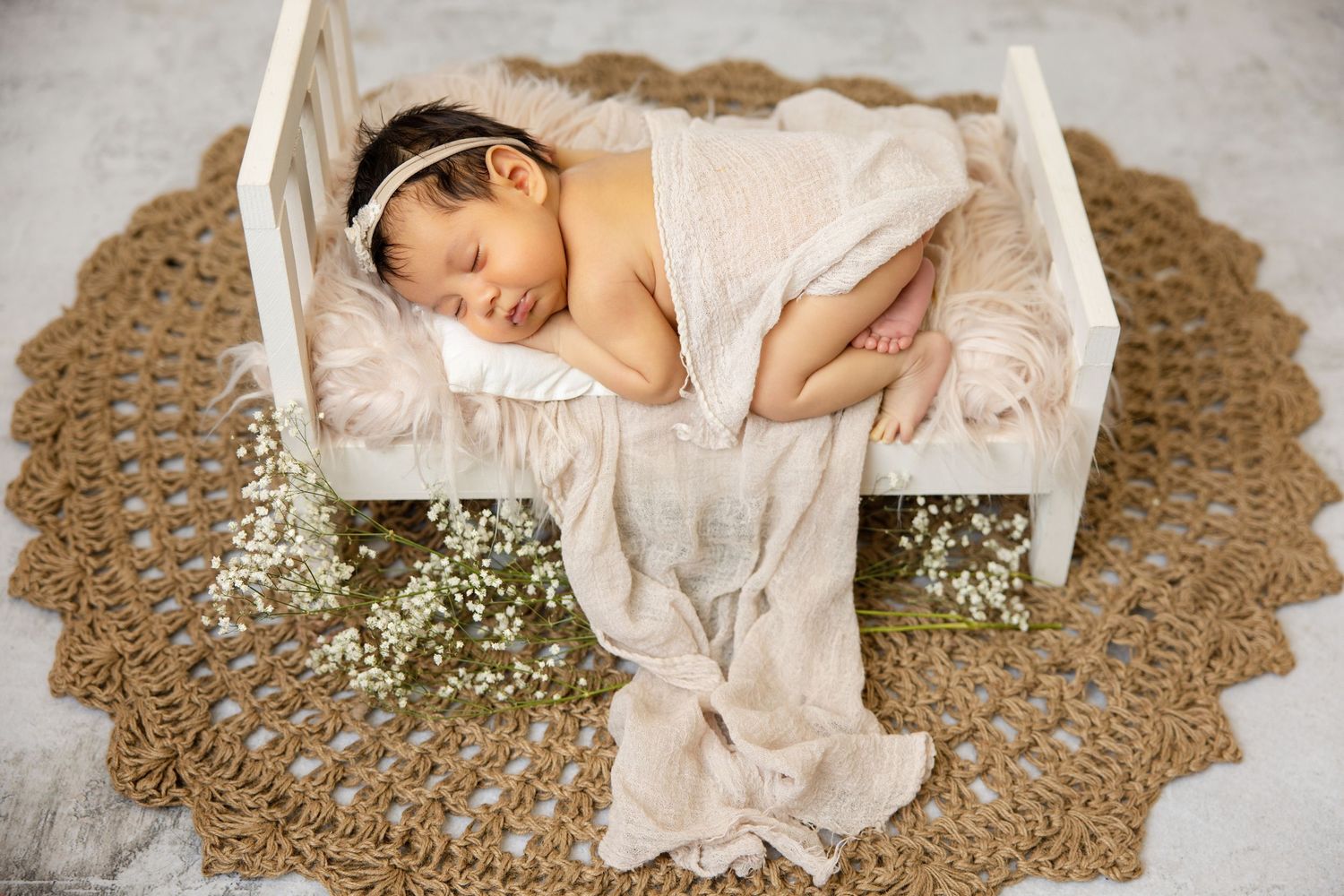 Newborn baby sleeping peacefully on a tiny white bed with floral decor on a woven rug.
