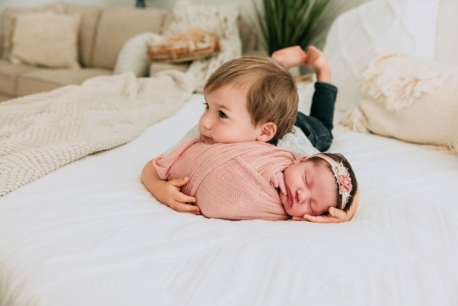 Toddler sibling lovingly rests chin on swaddled newborn baby in pink wrap on white bed.