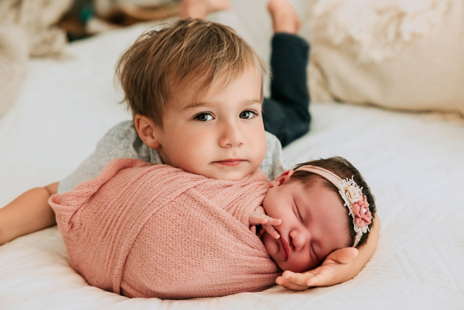 Toddler boy resting chin on swaddled newborn baby girl wearing pink floral headband on white bed.