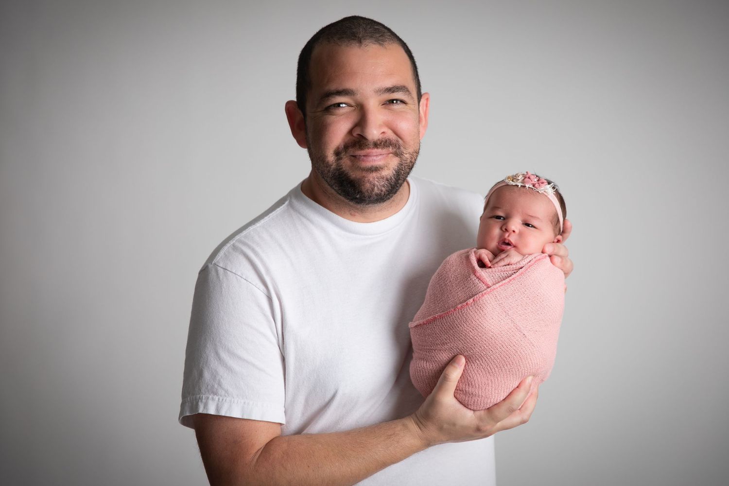A smiling man in a white t-shirt holds a swaddled newborn baby girl wearing a pink floral headband.