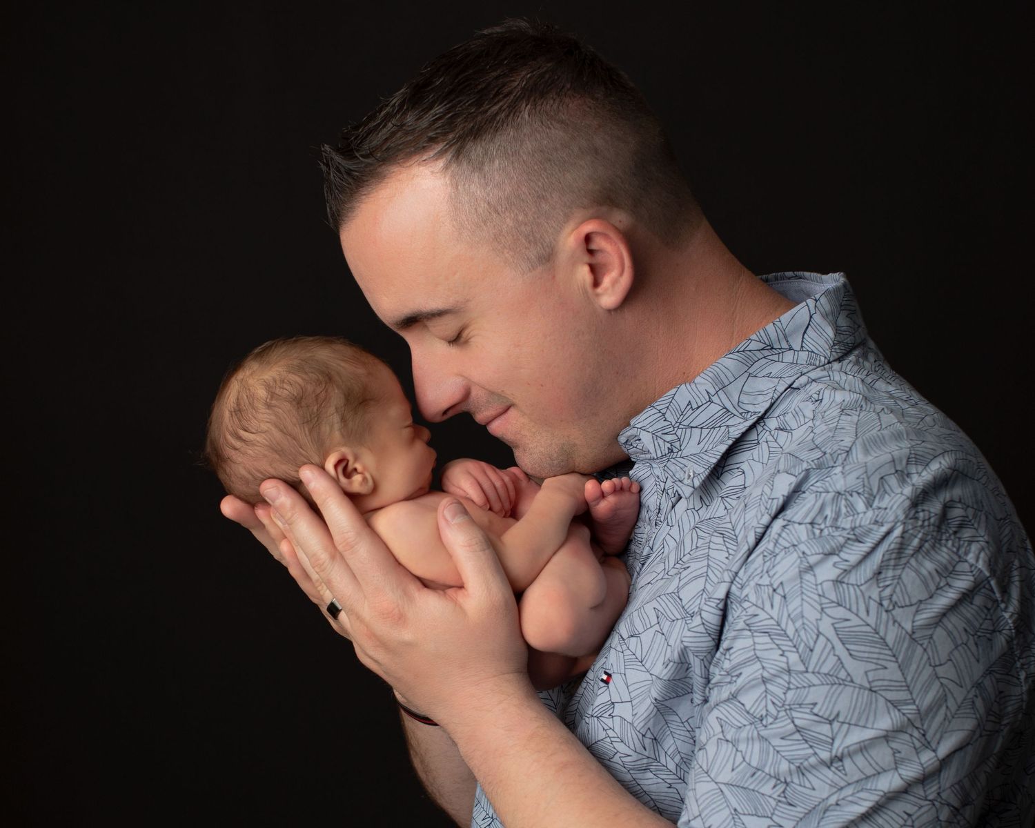 A father tenderly holds his newborn baby close to his face against a dark studio background.