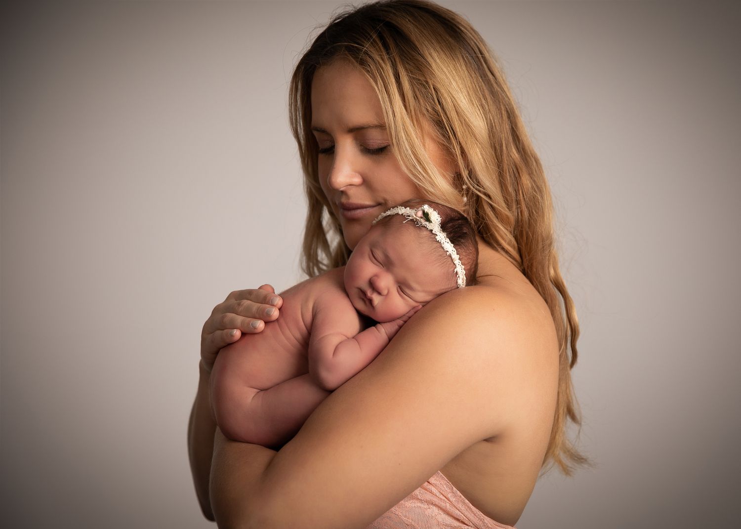 A mother tenderly holds her sleeping newborn baby against her chest in a soft studio portrait.
