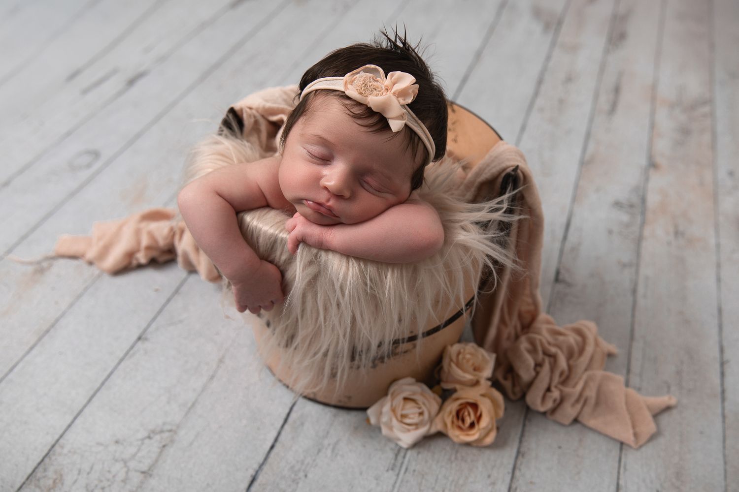 Sleeping newborn baby in a wooden bucket with fur, surrounded by roses, wearing a floral headband on a gray wood floor.
