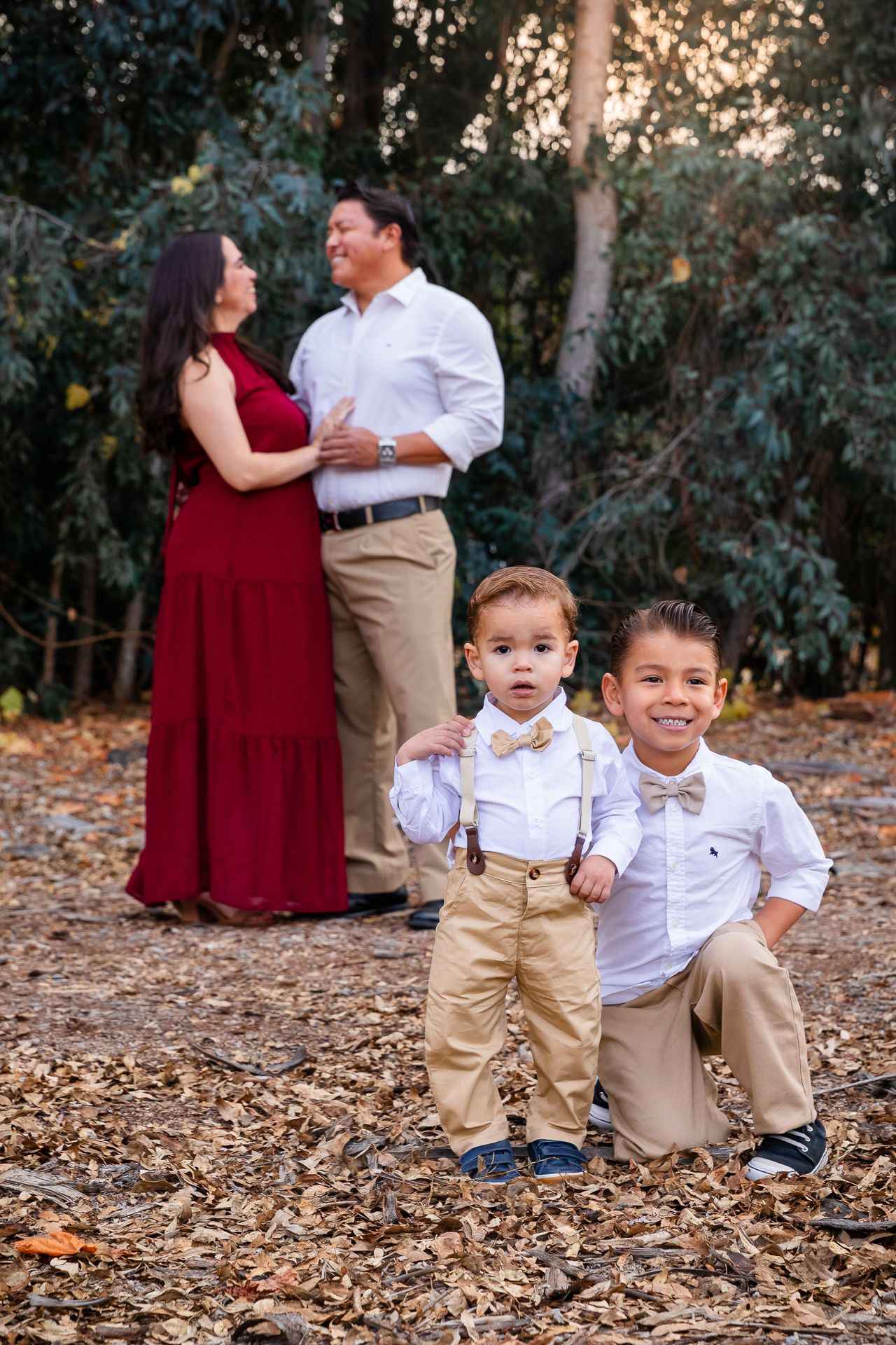 Two young boys in suspenders pose while parents embrace romantically among autumn leaves behind them.
