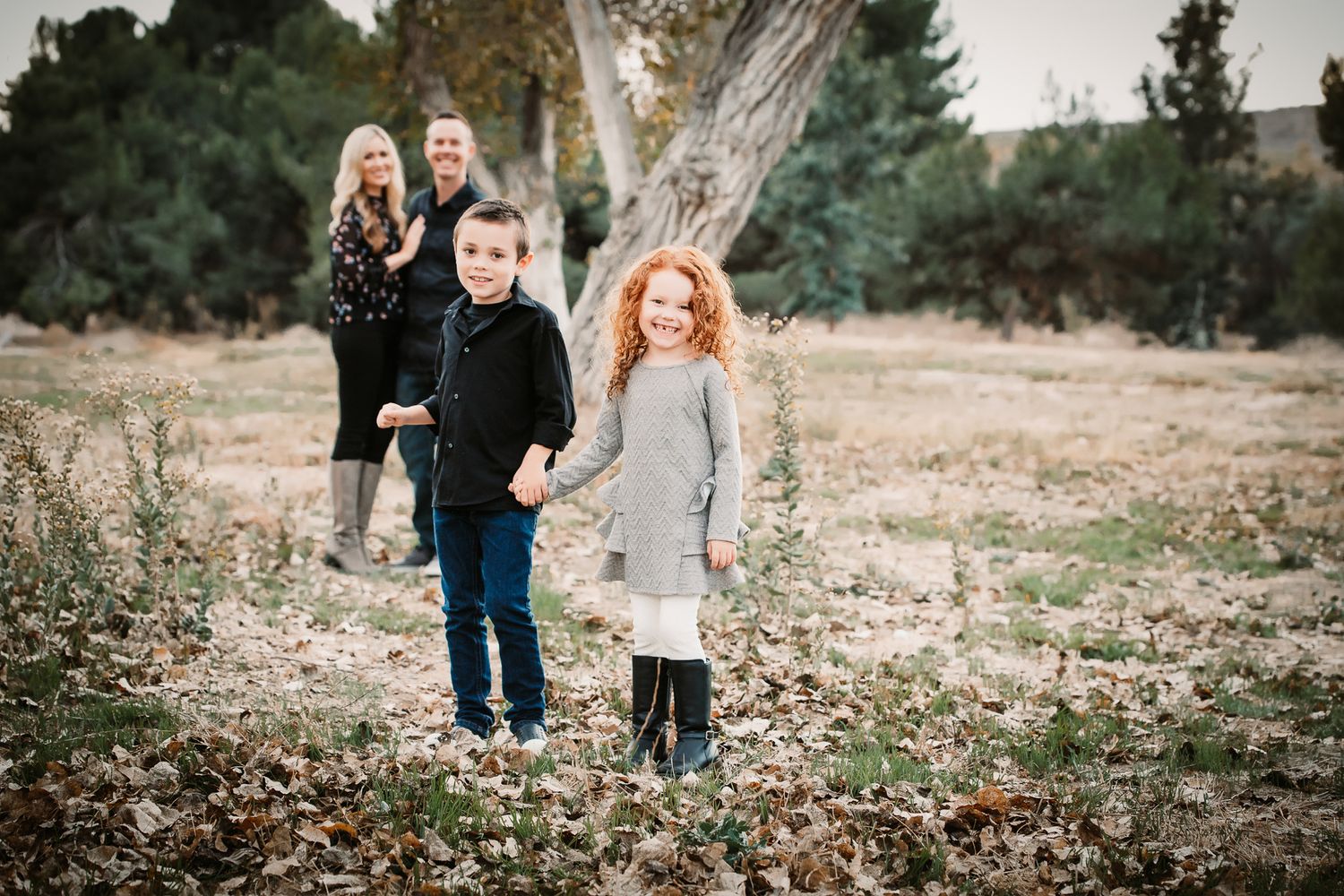 Two red-haired siblings hold hands in a leaf-covered field while parents stand behind them.