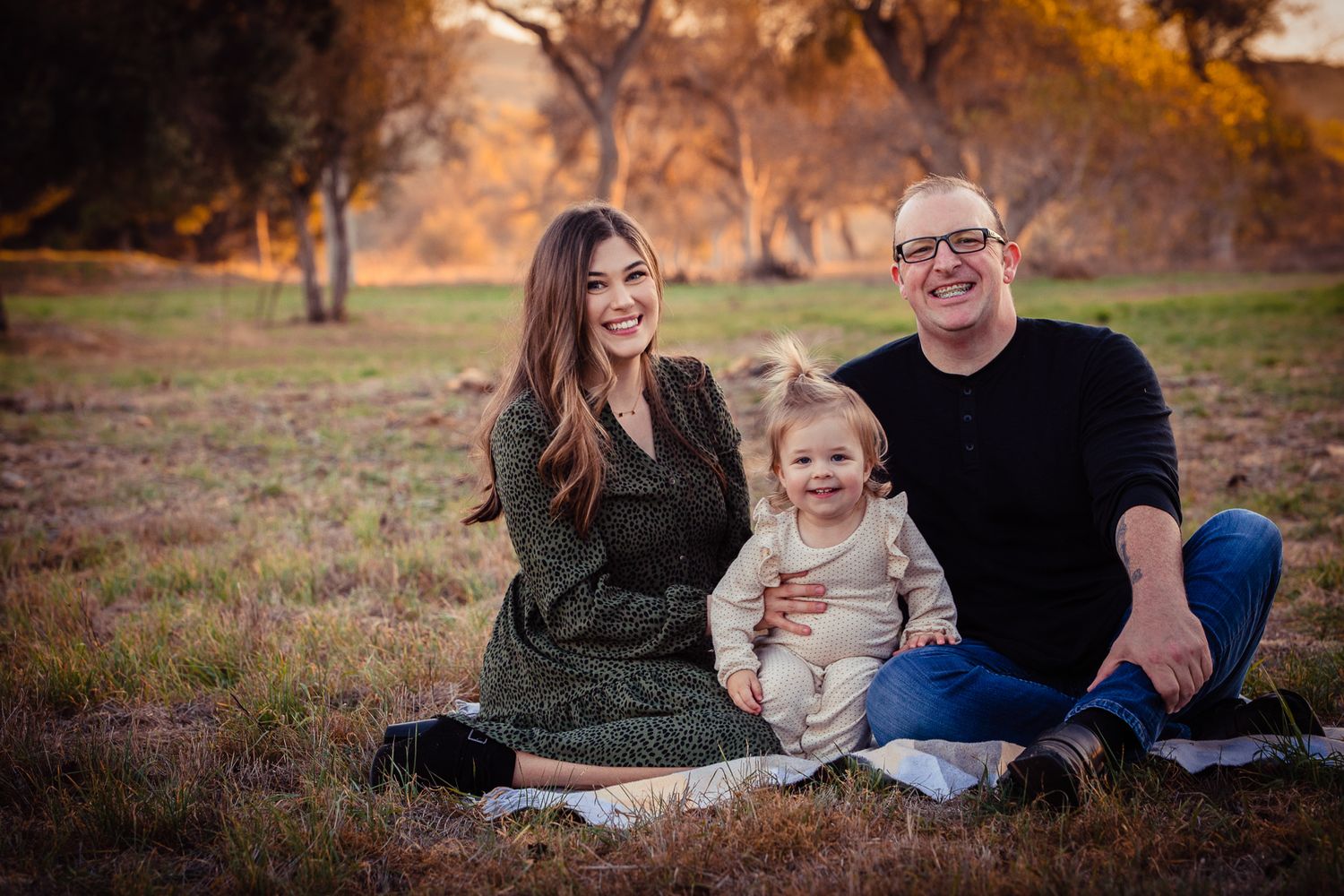Young family of three sitting on grass in park at sunset, smiling with toddler in Bonsall, near Fallbrook, & Temecula