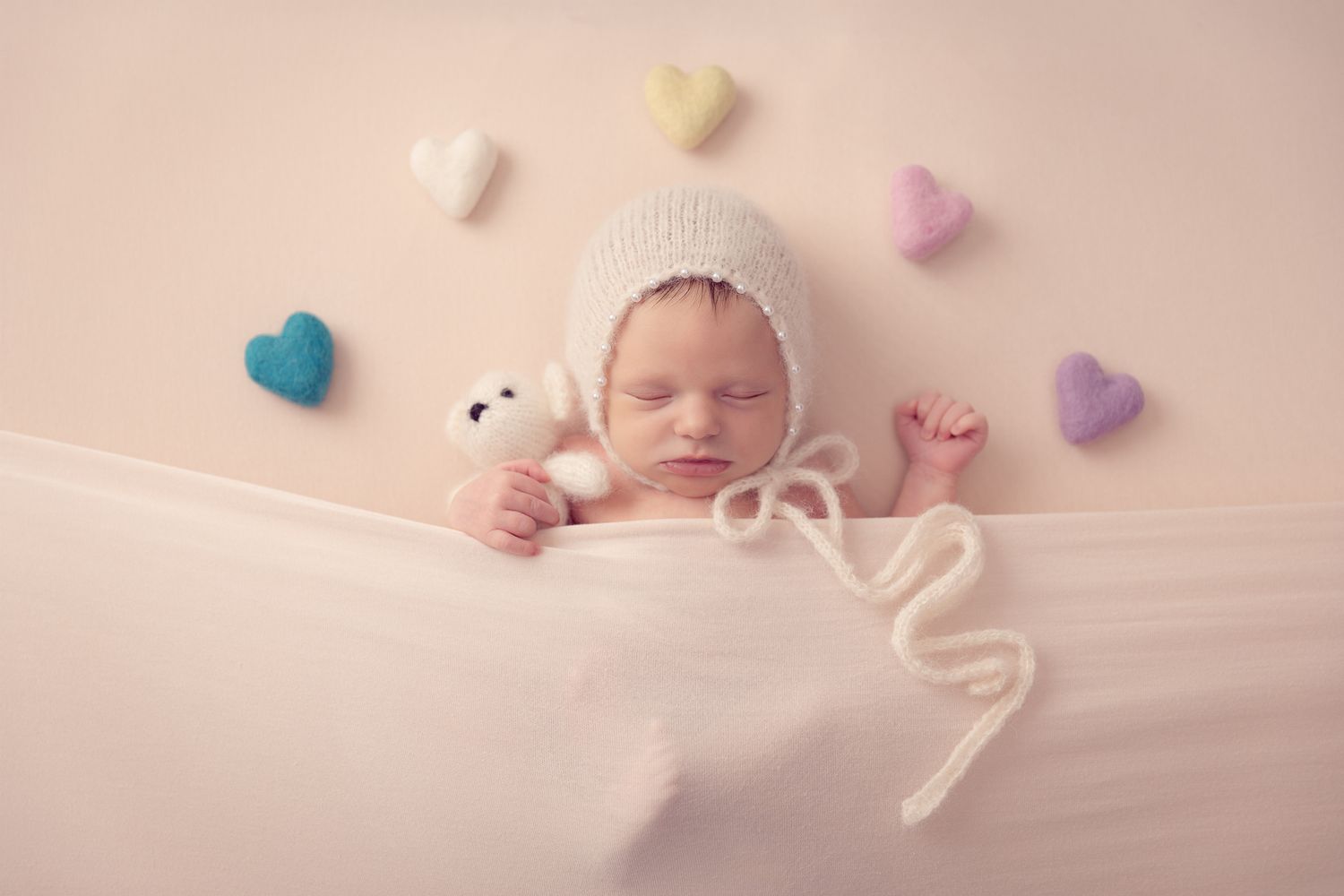 Sleeping newborn in white knit bonnet holds teddy bear surrounded by colorful felt hearts on pink backdrop.